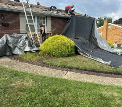 Roof replacement process with protective tarps in place to safeguard landscaping and property during work. Roof tear off in progress with crew and tarp protection setup on residential home in Pittsburgh PA